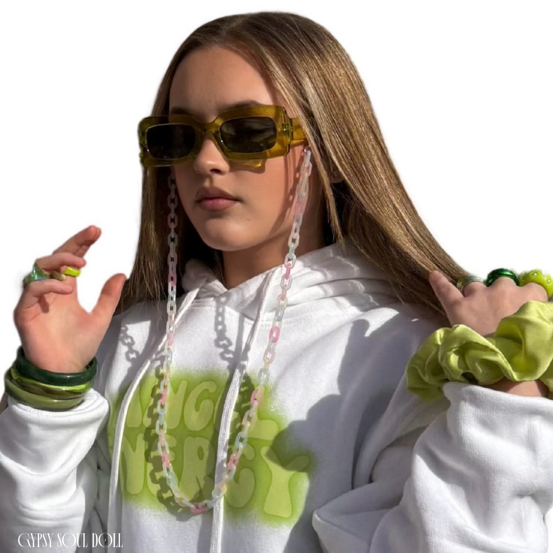 Model wearing olive green rectangular sunglasses attached to a pink, white, and green pastel acrylic link lanyard chain, styled with a white graphic hoodie, green wrist stack, and a lime green scrunchie.