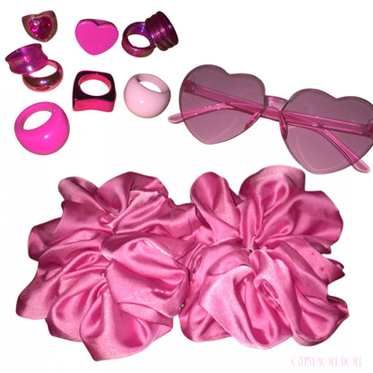 A curated Y2K candy pink accessory bundle featuring two oversized pink satin scrunchies, a pair of frameless translucent heart-shaped sunglasses, and an assortment of retro resin rings including 3D puffy hearts, metallic pink bands, and chunky acrylic domes on a plain white background.