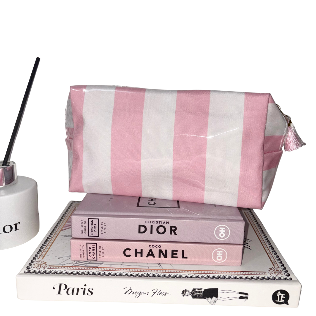 Candy Stripe cosmetic bag in glossy pink and white stripes featuring a tassel zip pull, displayed on top of decorative fashion books next to a room diffuser.