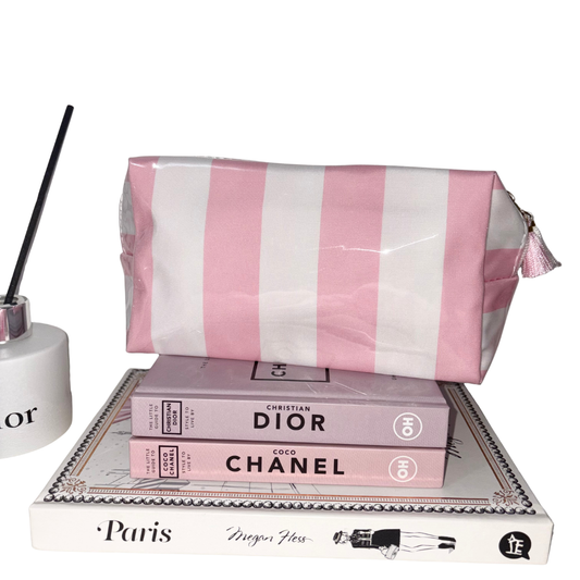 Candy Stripe cosmetic bag in glossy pink and white stripes featuring a tassel zip pull, displayed on top of decorative fashion books next to a room diffuser.