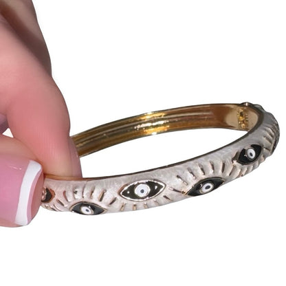 Side profile view of a hand with a white tipped French manicure holding a polished gold bangle bracelet featuring a pearlescent white enamel coating and striking black and white evil eye motifs with golden lashes.