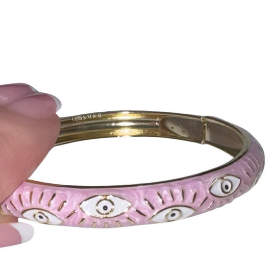 Side angle view of a hand holding a polished gold bangle bracelet featuring a pastel pink enamel coating with white and gold evil eye protective motifs.
