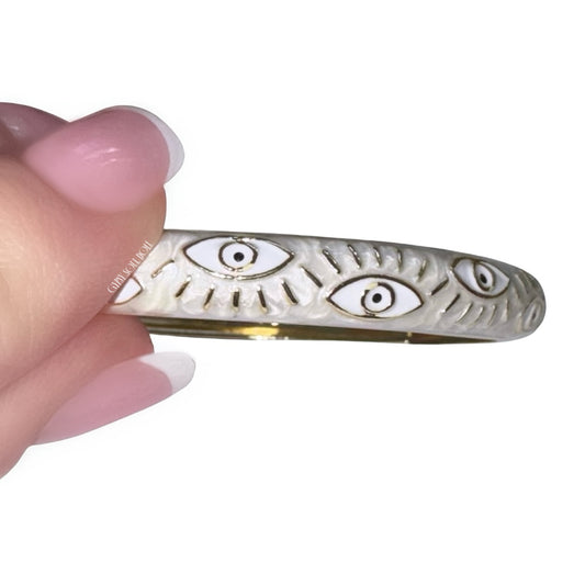 Side angle view of fingers with a pink french manicure holding a polished gold bangle bracelet featuring a creamy white enamel coating with white and gold evil eye protective motifs.
