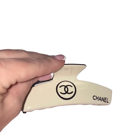 A hand with a classic french manicure holding a curved cream beige hair claw clip featuring black designer block text and an interlocking circle logo design against a white background.