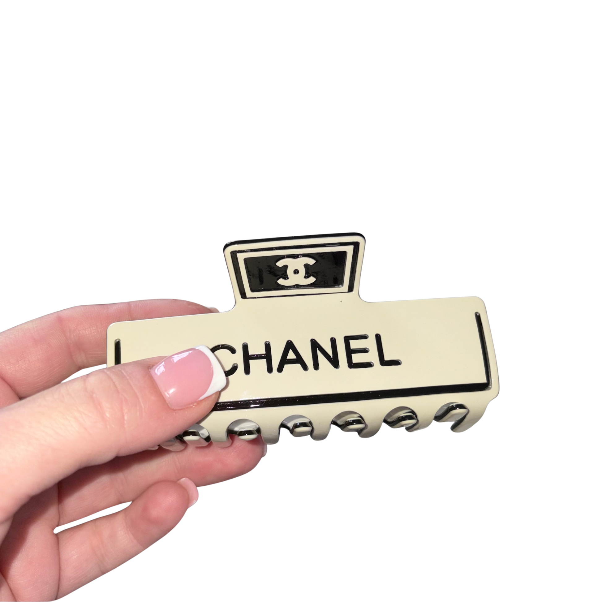 A hand with a French manicure holds a cream-coloured Chanel hair claw clip with black lettering and logo details against a plain white background.