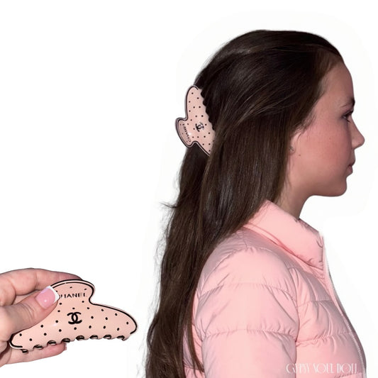 A pink claw hair clip with black polka dots and a Chanel logo, shown being held by a hand on the left and worn in a girl's hair on the right. A model in a pink puffer jacket is shown in profile against a white background.