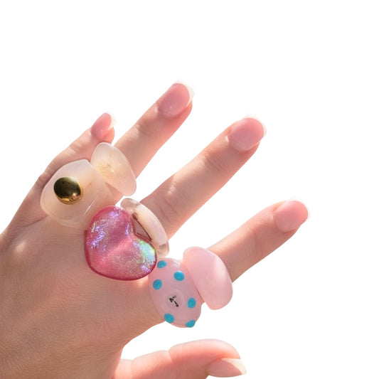 Close-up of a hand with a classic French manicure showcasing a stacked collection of translucent lemon yellow, blue polka dot, and glittery pink heart chunky acrylic statement rings.