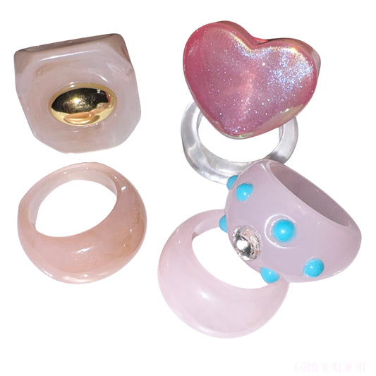 Six decorative rings arranged on a plain white background: a translucent pale pink square ring with a gold-toned center, a pink glitter heart-shaped ring on a clear band, a translucent pale pink band ring, a pale pink ring with bright blue dots and a clear rhinestone, and a matching translucent pale pink ring shown from the back.