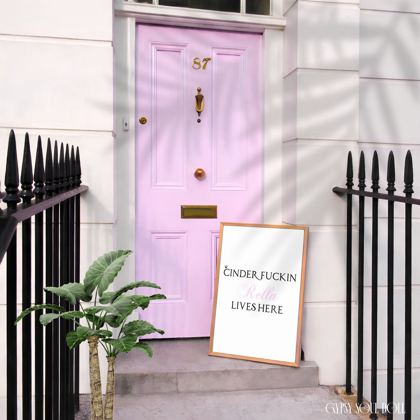 A framed art print with the text 'CINDER, FUCKIN' DONT LET THEM LIVES HERE' in black and pink lettering on a white background, leaning on the steps next to a light pink front door.
