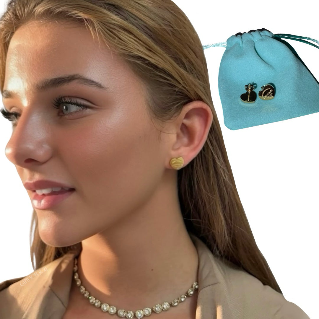 A side profile view of a young woman wearing small gold heart-shaped stud earrings and a gold bezel tennis necklace, pictured next to a turquoise blue drawstring pouch containing a pair of silver heart studs.