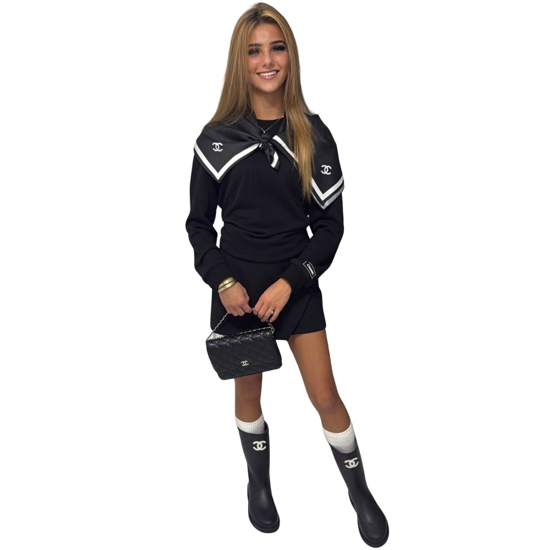 A woman modeling a black Chanel long-sleeve mini dress with white geometric shoulder stripes and double-C logos on the chest and sleeve. She is wearing black rain boots with white socks, holding a black quilted Chanel handbag, against a solid white background.