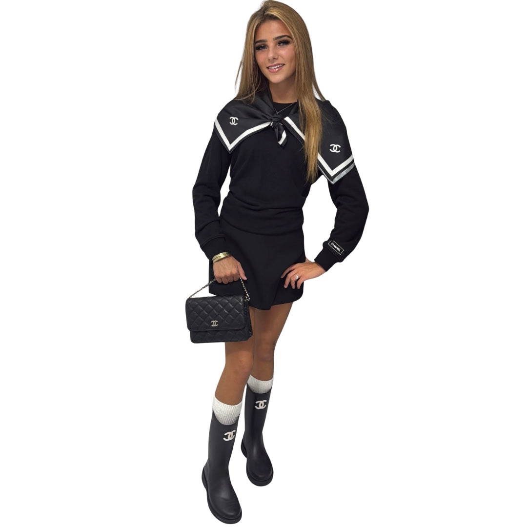 A woman modelling a black Chanel mini dress with white stripes and double-C logos on the chest and sleeve, paired with white socks, black rain boots, and a black quilted Chanel handbag, set against a plain white background.