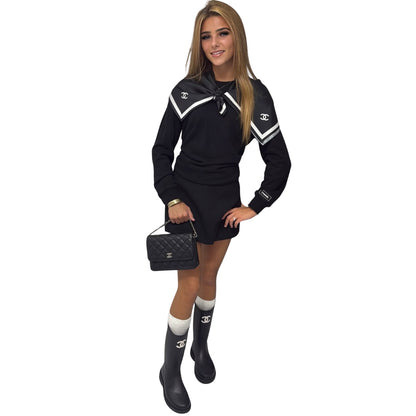 A woman modelling a black Chanel mini dress with white stripes and double-C logos on the chest and sleeve, paired with white socks, black rain boots, and a black quilted Chanel handbag, set against a plain white background.