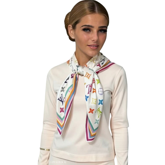 A model wearing a cream loungewear outfit, styled with a white scarf featuring a colourful, multicoloured monogram pattern and striped borders. The scarf displays the Louis Vuitton monogram motif.