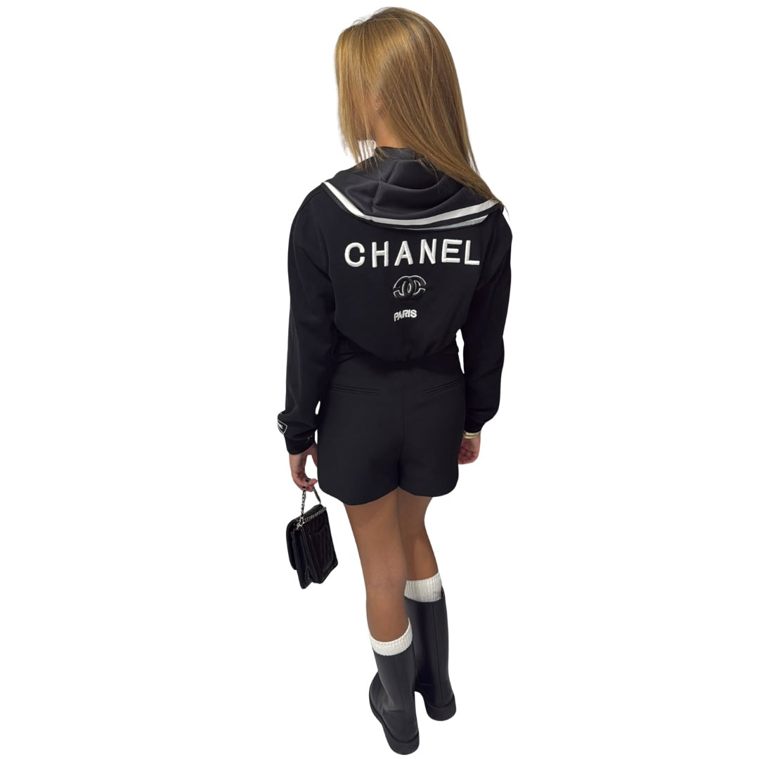 A woman shown from the back wearing a black Chanel top and matching classy yacht shorts with white lettering and stripes, paired with black rain boots and holding a small black handbag against a plain white background.