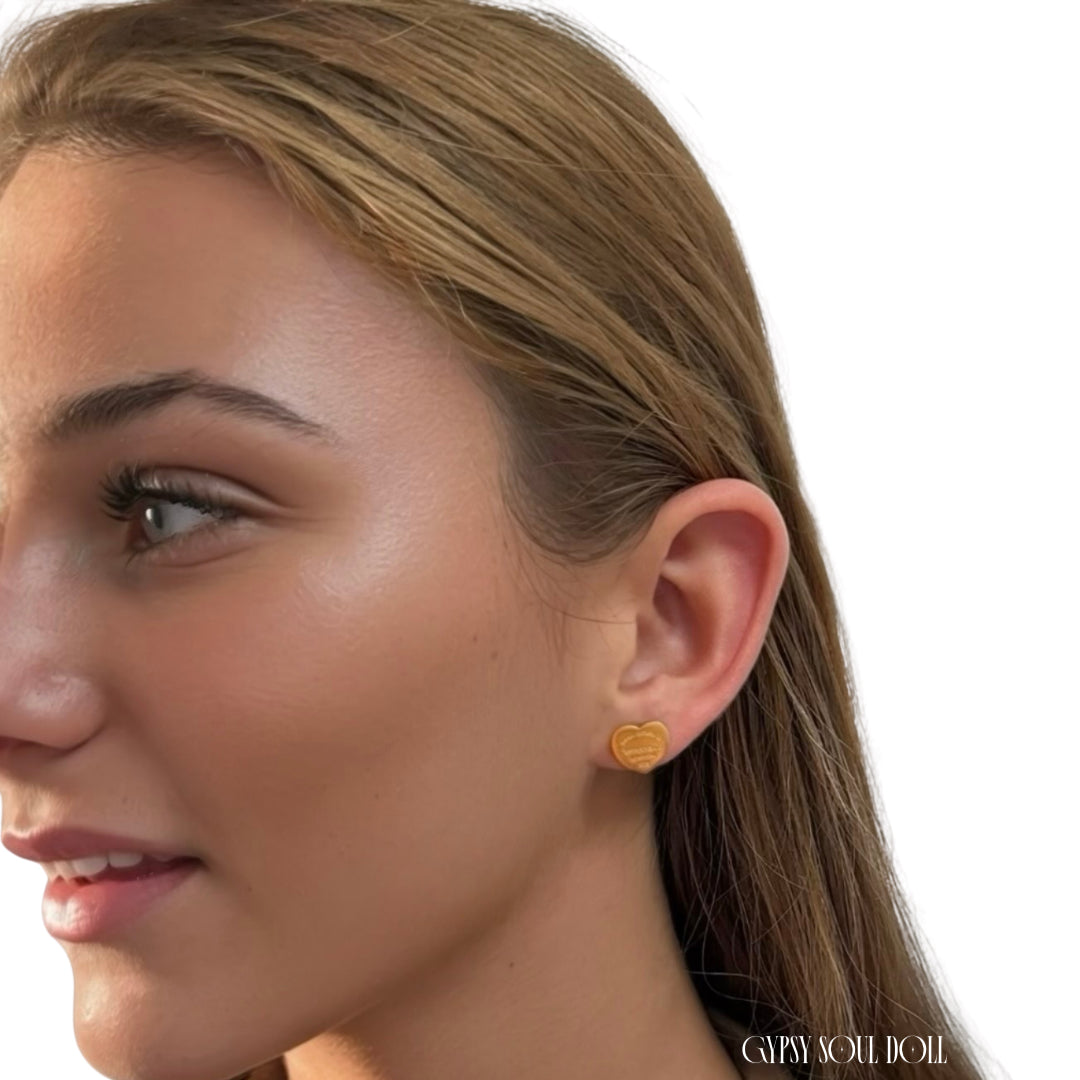 A close-up side profile of a woman wearing a small gold heart-shaped designer-inspired stud earring. The brand name 'GYPSY SOUL  DOLL' is visible in the bottom right corner.
