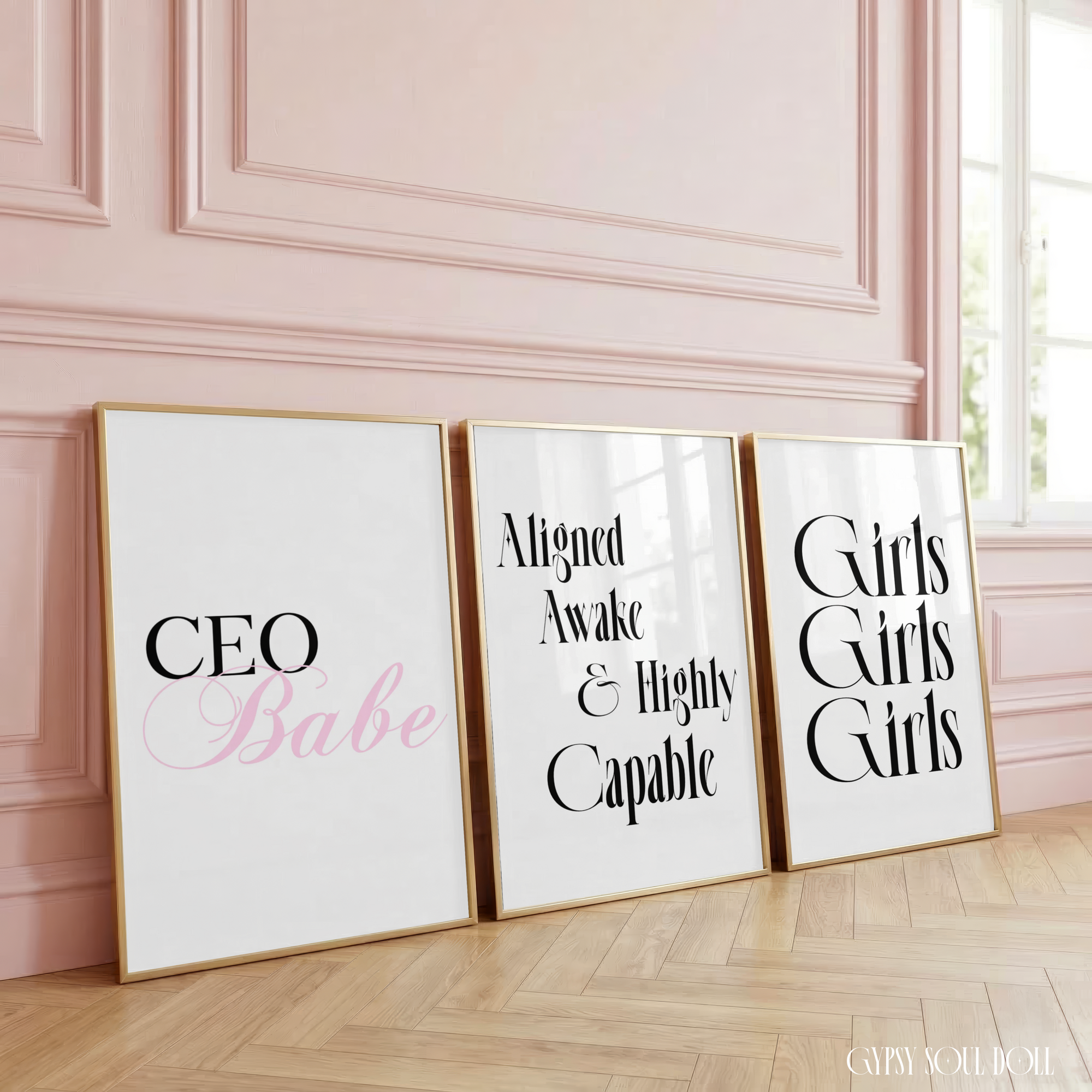The Corporate Doll three-piece minimalist typography print set is beautifully styled in sleek gold frames, leaning against a soft pink panelled luxury wall on light herringbone wood flooring.
