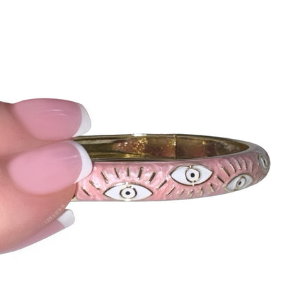 Side profile view of a hand with a soft pink French manicure holding a polished gold bangle bracelet featuring a textured pink enamel coating and white and black evil eye motifs with golden lashes by Gypsy Soul Doll.