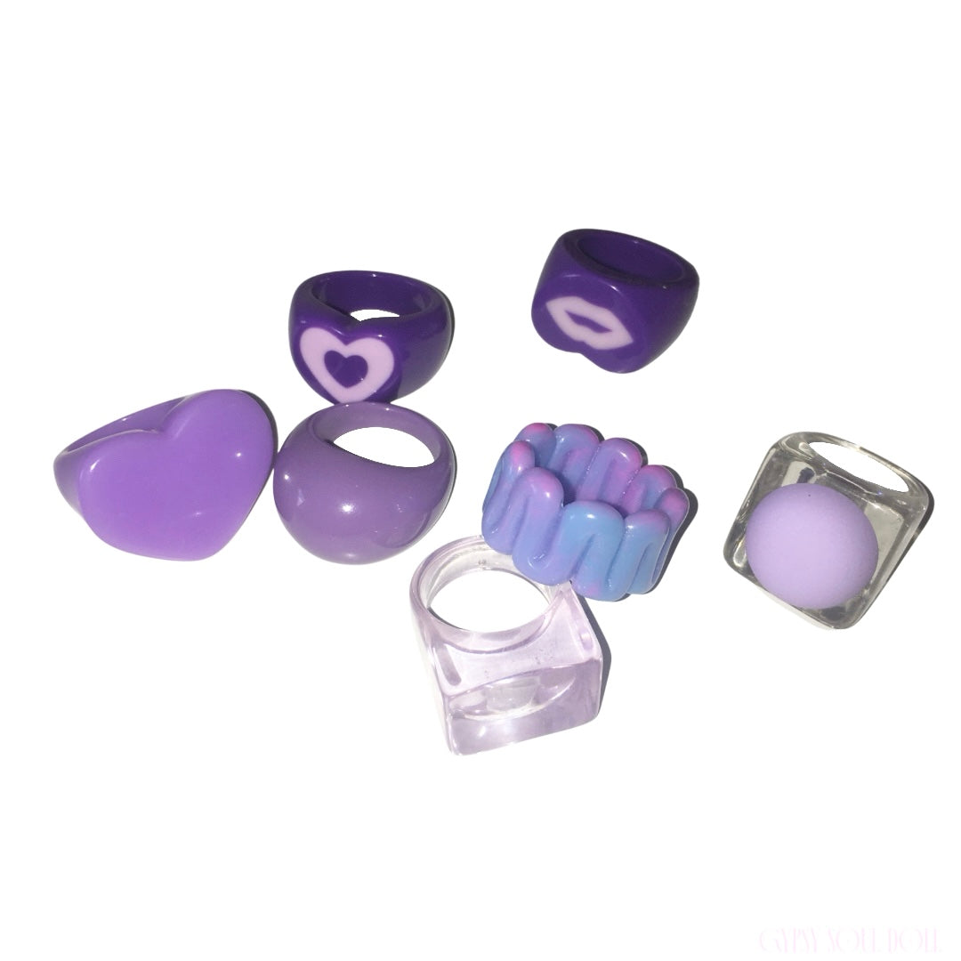 A set of seven purple silicone acrylic rings displayed on a clean white background. The collection includes a heart-shaped bead, a smooth oval bead, a ring with a  heart, a ring with a white lips motif, a textured blue-and-purple bead, a clear square bead containing a purple sphere, and a clear square ring.