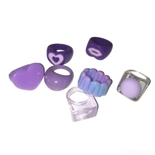 A set of seven purple silicone acrylic rings displayed on a clean white background. The collection includes a heart-shaped bead, a smooth oval bead, a ring with a  heart, a ring with a white lips motif, a textured blue-and-purple bead, a clear square bead containing a purple sphere, and a clear square ring.