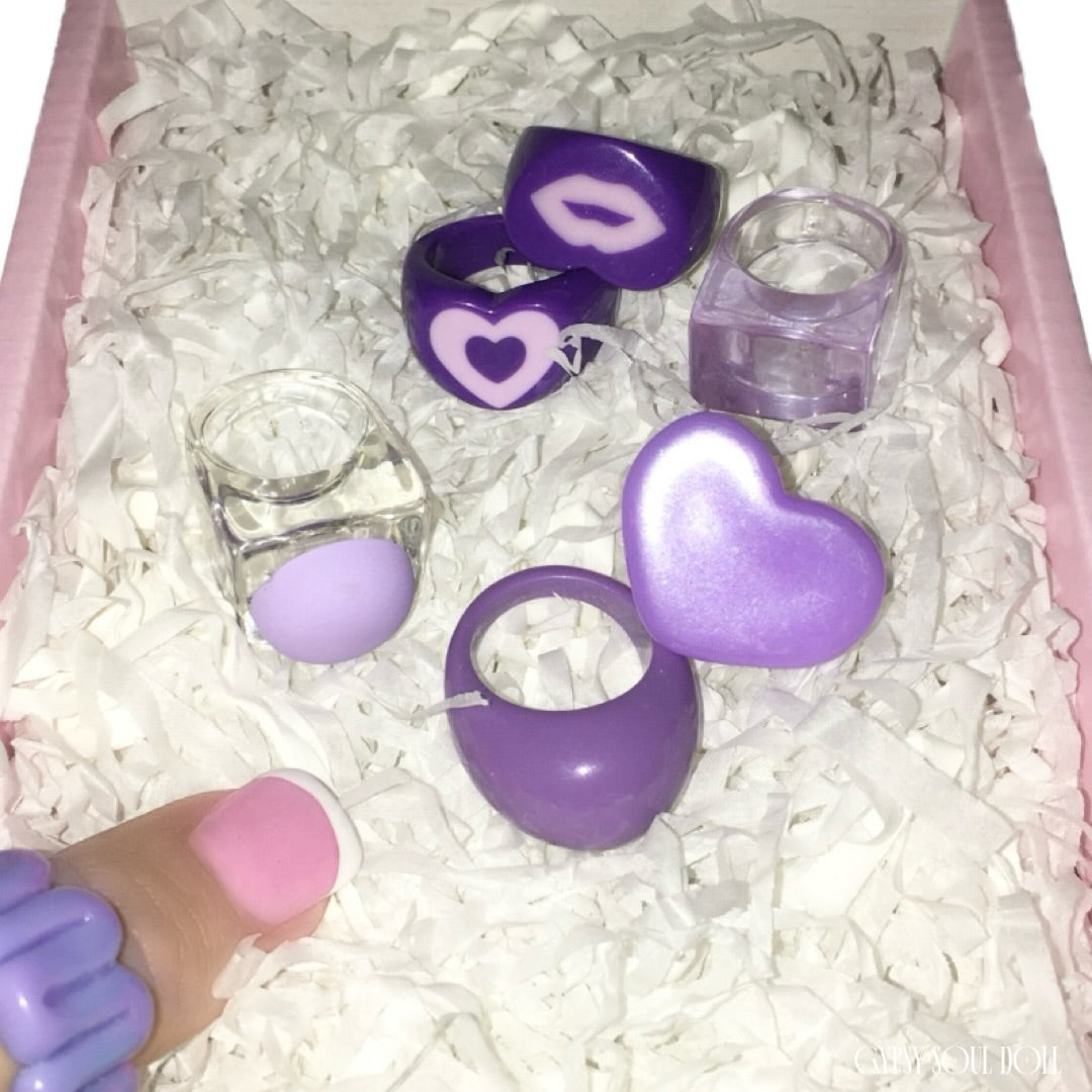 Top-down view of six chunky Y2K festival style acrylic and resin rings in shades of purple and transparent clear, featuring moulded lip and heart designs, nestled inside a pink Gypsy Soul Doll boutique presentation box with white crinkle paper.
