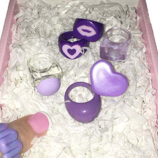 Top-down view of six chunky Y2K festival style acrylic and resin rings in shades of purple and transparent clear, featuring moulded lip and heart designs, nestled inside a pink Gypsy Soul Doll boutique presentation box with white crinkle paper.