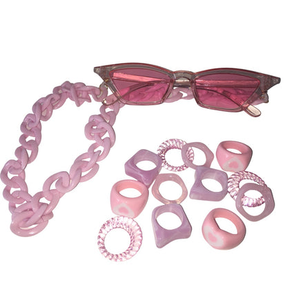 Flat lay view of the Gypsy Soul Doll Cyber Candy festival combo, featuring translucent pink cat-eye fashion glasses with a thick pink acrylic link chain lanyard next to a scattered assortment of pink resin heart rings, geometric bands, and clear spiral rings.