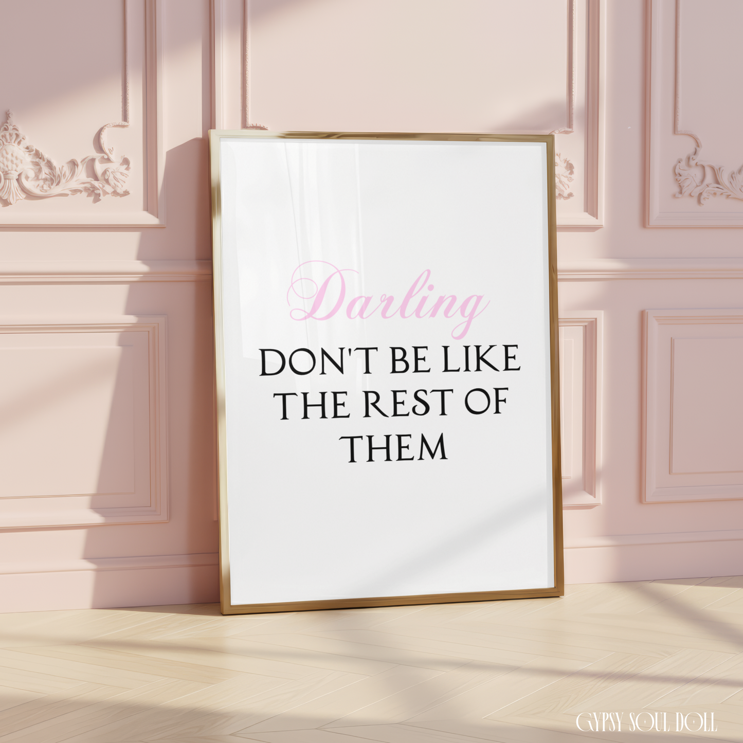 Gold-framed typography print leaning on light wood flooring against a dusty pink panelled wall, reading Darling Don't Be Like The Rest Of Them with Darling in pink script.