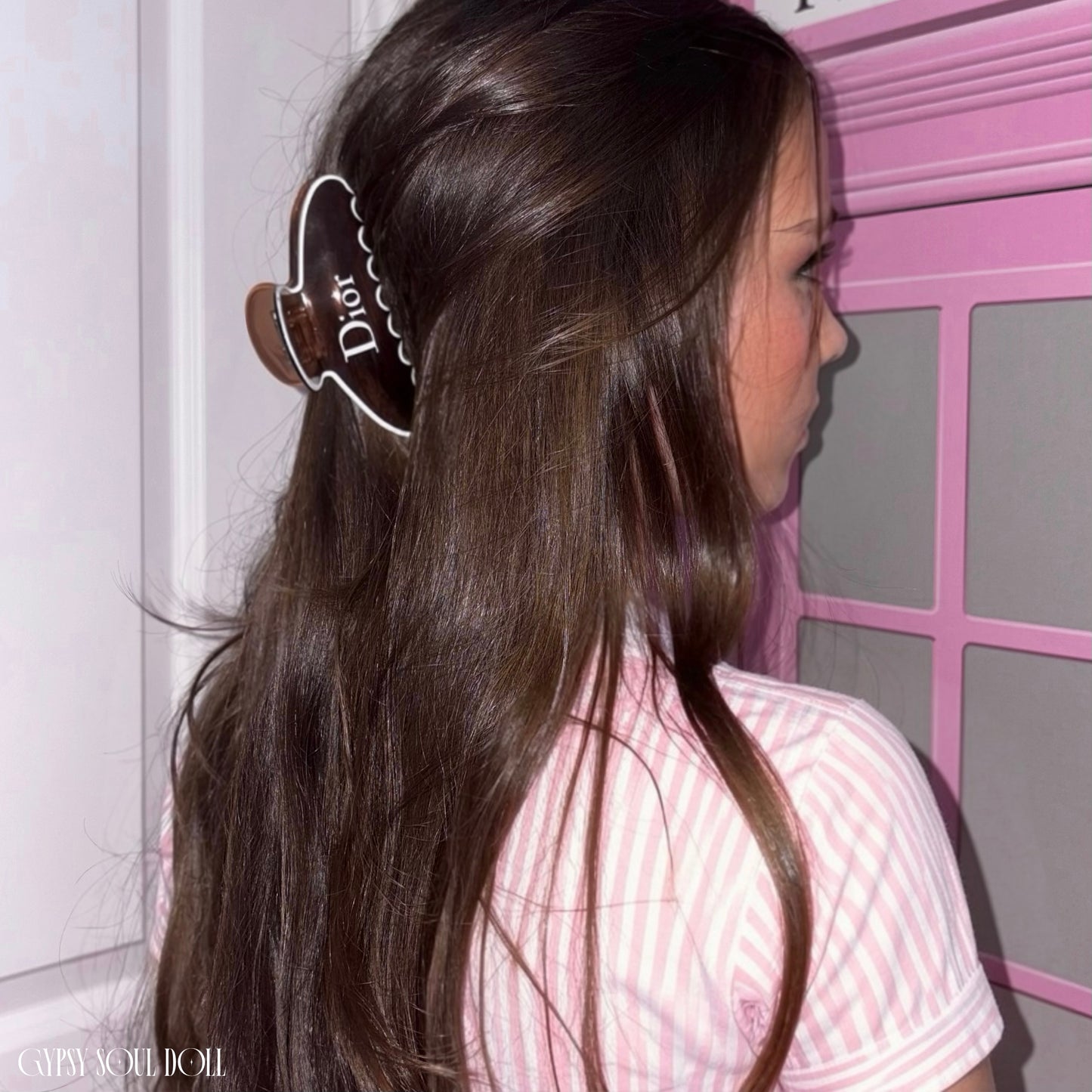 Rear view of a model with long glossy brown hair styled in a half-up look secured by a Gypsy Soul Doll tortoiseshell claw clip featuring white script text that reads ‘Dior’ wearing a pink and white striped shirt.