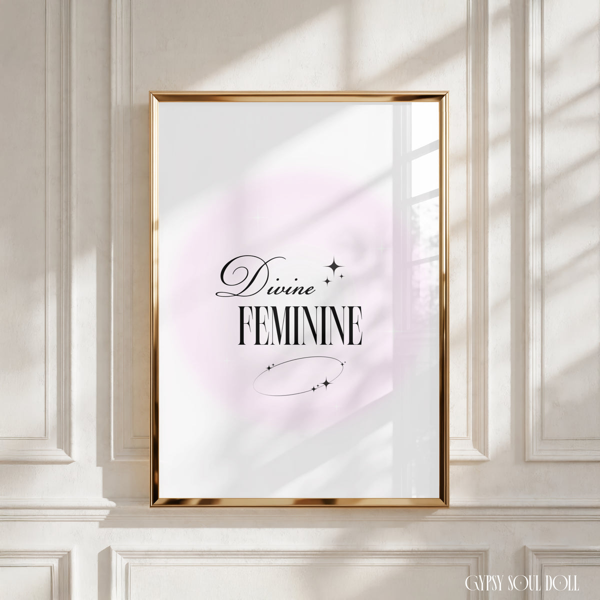 A framed art print with the words 'Divine FEMININE' in black script and serif lettering, set against a soft pink circular backdrop on a white background. The gold frame is mounted on a white panelled wall, with realistic window-shadow light cast across the scene by Gypsy Soul Doll.
