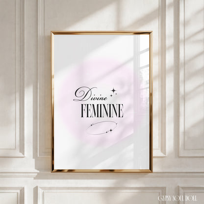A framed art print with the words 'Divine FEMININE' in black script and serif lettering, set against a soft pink circular backdrop on a white background. The gold frame is mounted on a white panelled wall, with realistic window-shadow light cast across the scene by Gypsy Soul Doll.