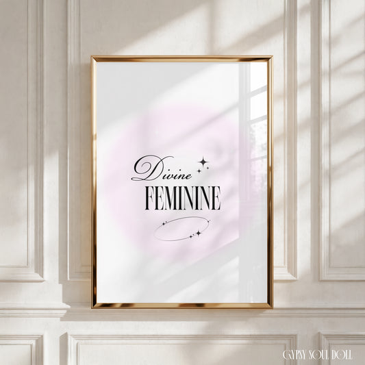 A framed art print with the words 'Divine FEMININE' in black script and serif lettering, set against a soft pink circular backdrop on a white background. The gold frame is mounted on a white panelled wall, with realistic window-shadow light cast across the scene by Gypsy Soul Doll.