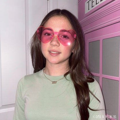 Close-up portrait of a smiling model with long dark brown hair wearing a delicate gold-plated link chain necklace with a 222 angel number pendant, paired with pink frameless heart-shaped sunglasses.