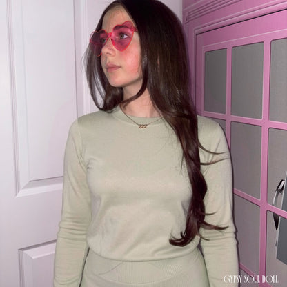 Model with long dark hair looking to the side while wearing a sage green top and the Gypsy Soul Doll dainty gold link chain necklace featuring a 222 angel number block charm and pink heart fashion glasses.