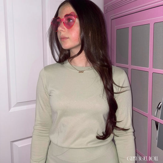 Model with long dark hair looking to the side while wearing a sage green top and the Gypsy Soul Doll dainty gold link chain necklace featuring a 222 angel number block charm and pink heart fashion glasses.