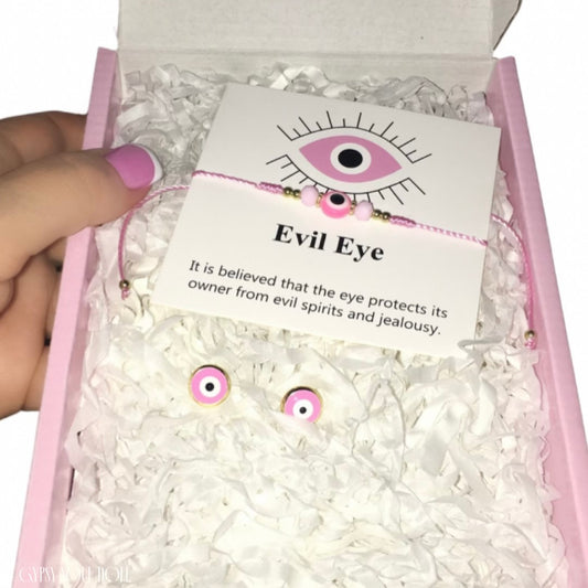 Flat lay view of the Gypsy Soul Doll Pink Evil Eye protective jewellery set, featuring an adjustable pink cord multi-wear bracelet or anklet wrapped around a white display card with eye meaning  text, and two matching round pink enamel stud earrings resting in a pink presentation box with white shredded tissue paper.