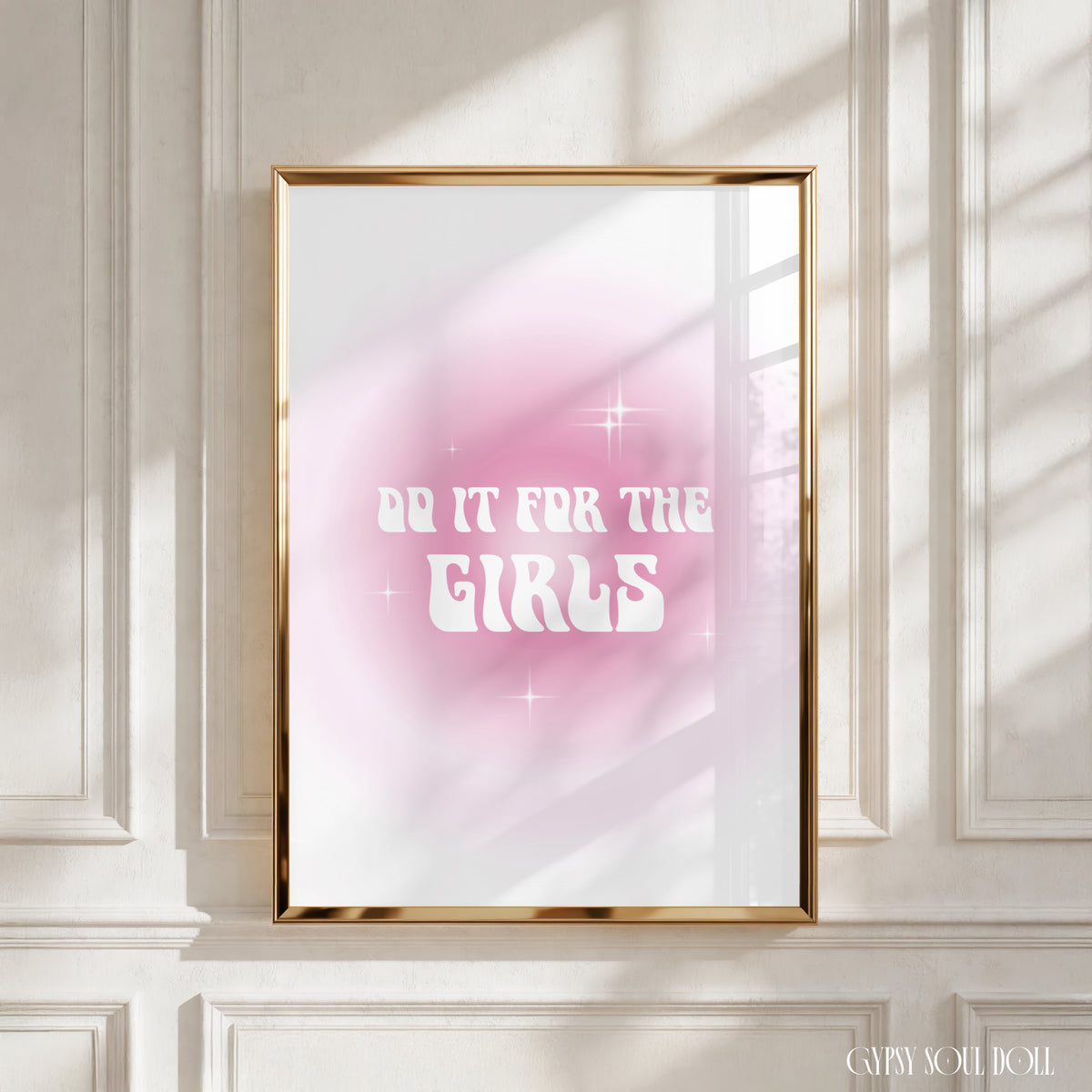 A framed art print with the text 'DO IT FOR THE GIRLS' in white against a pink and white gradient background, displayed on a white panelled wall with soft window shadows by Gypsy Soul Doll.