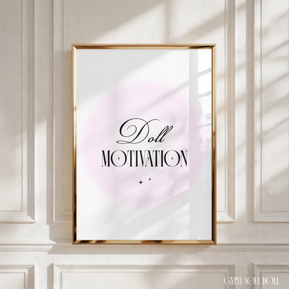 A framed typographic wall art print with the words 'Doll MOTIVATION' in black script and serif lettering over a soft pale pink circular backdrop, displayed against an off-white panelled wall with warm window light casting shadows by Gypsy Soul Doll.