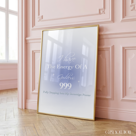 A vertical framed art print with a gold frame leans against a pink paneled wall on a wooden floor. The print features a soft gray and white gradient background with elegant white text that reads, 'I have The Energy Of A Goddess 999 Fully Stepping Into My Screen Sign Power.