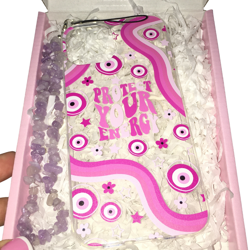 A clear phone case with a pink and white graphic design featuring the text 'PROTECT YOUR ENERGY' and eye motifs, presented inside a pink gift box with white paper shred and purple crystals. A hand is holding the left side of the box.