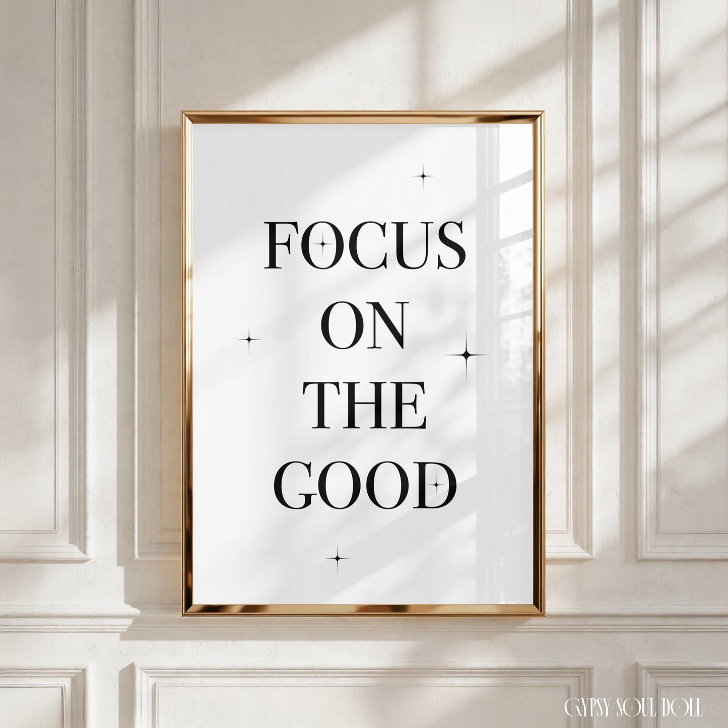 A framed typographic wall art print with the words “FOCUS ON THE GOOD” in black serif lettering on a white background, set in a thin gold frame and displayed against a cream panelled wall with soft window shadows.