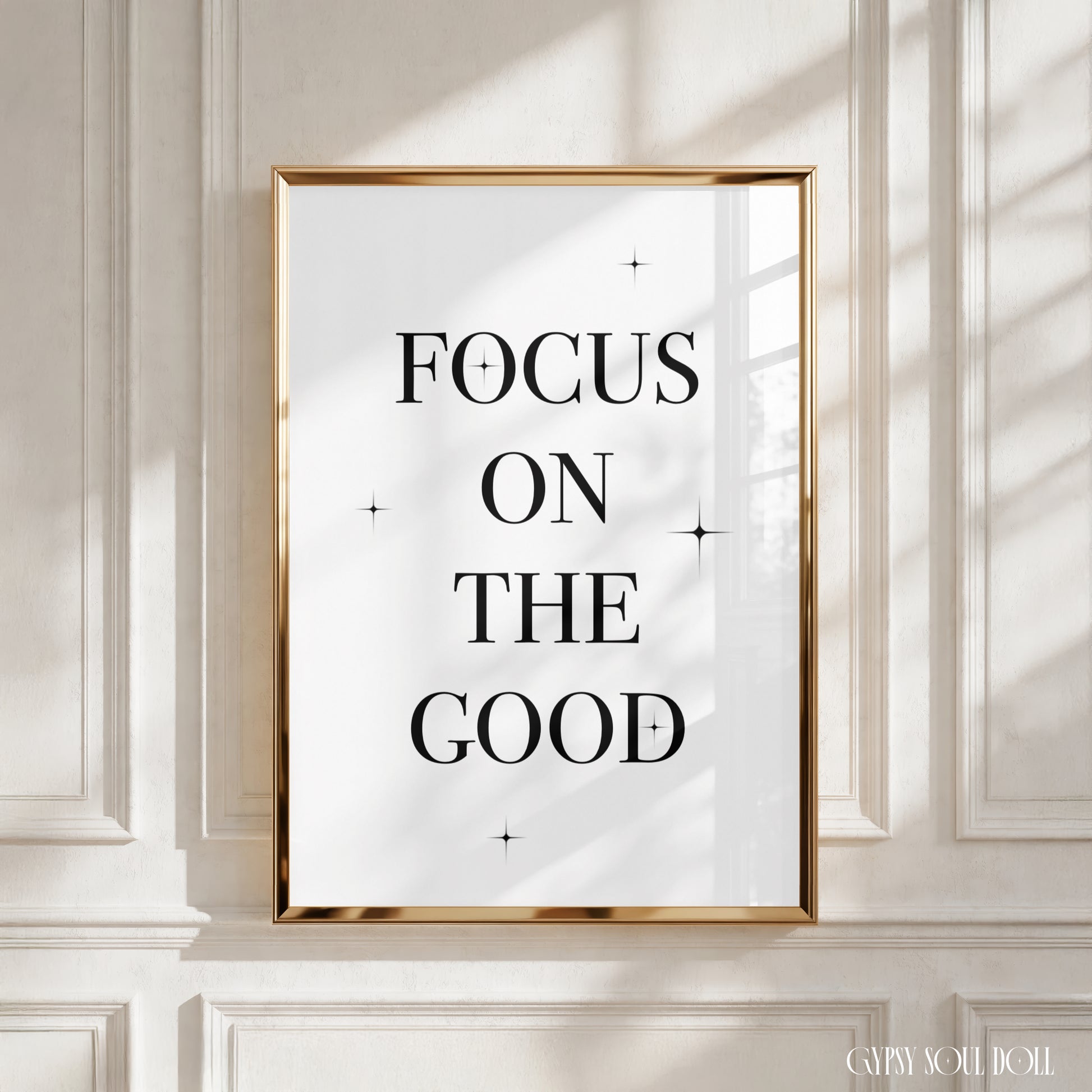 A framed typographic wall art print with the words “FOCUS ON THE GOOD” in black serif lettering on a white background, set in a thin gold frame and displayed against a cream panelled wall with soft window shadows.