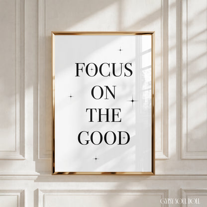A framed typographic wall art print with the words “FOCUS ON THE GOOD” in black serif lettering on a white background, set in a thin gold frame and displayed against a cream panelled wall with soft window shadows.