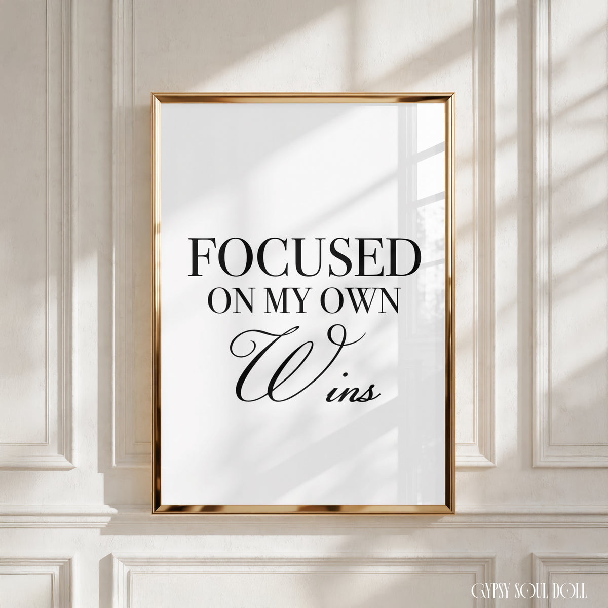 A framed typographic wall art print with the text 'FOCUSED ON MY OWN Wins' in black lettering on a white background, set in a thin gold frame against an off-white panelled wall with soft window shadows by Gypsy Soul Doll.