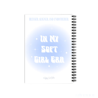A white spiral notebook with a light blue and white graphic design on the cover. The cover features the text 'BLESSED, ALIGNED, UNBOTHERED and In My Soft Girl Era by Gypsy Soul Doll.