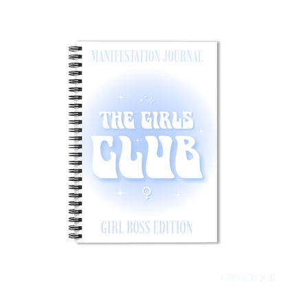 A white spiral-bound notebook with a black wire binding, featuring the text 'MANIFESTATION JOURNAL', 'THE GIRLS CLUB', and 'GIRL BOSS EDITION' in white and light blue on the cover by Gypsy Soul Doll.