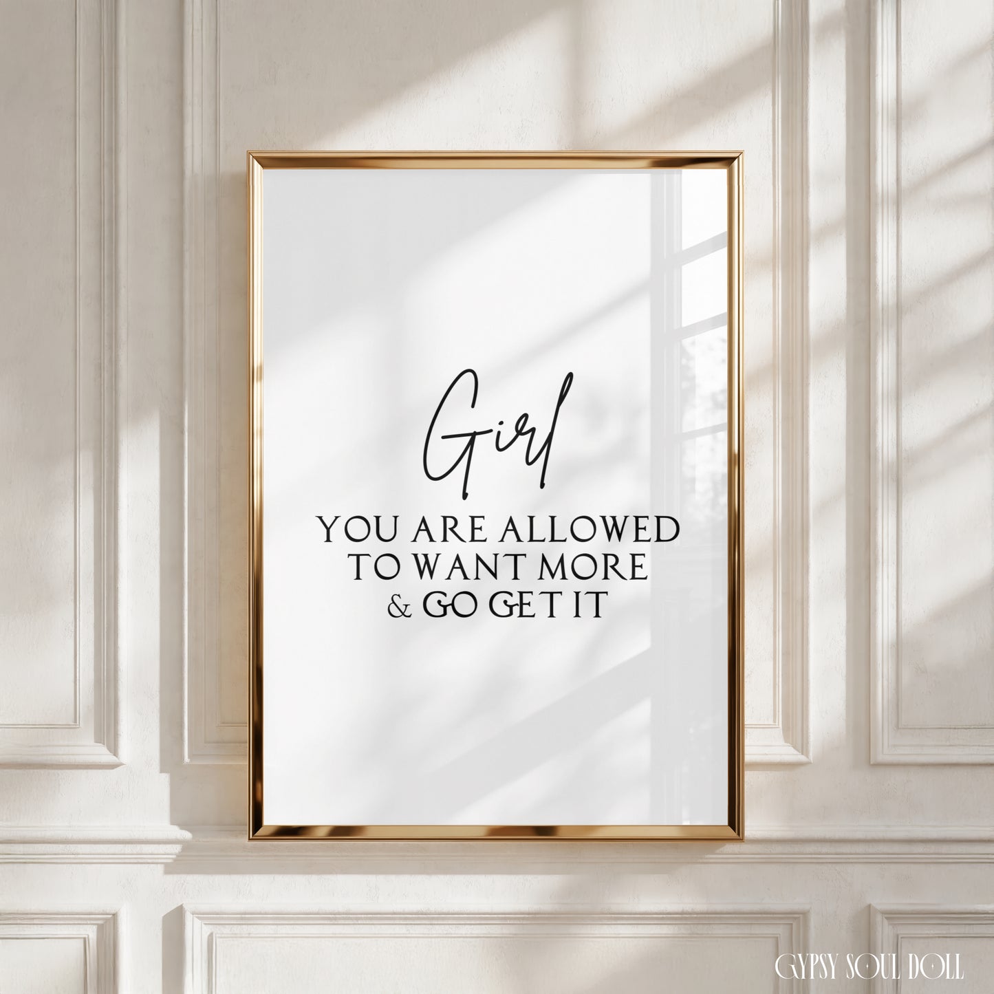 A framed typographic wall art print with a gold frame hangs on a white paneled wall. The print has black text reading “Girl YOU ARE ALLOWED TO WANT MORE & GO GET IT” on a white background.