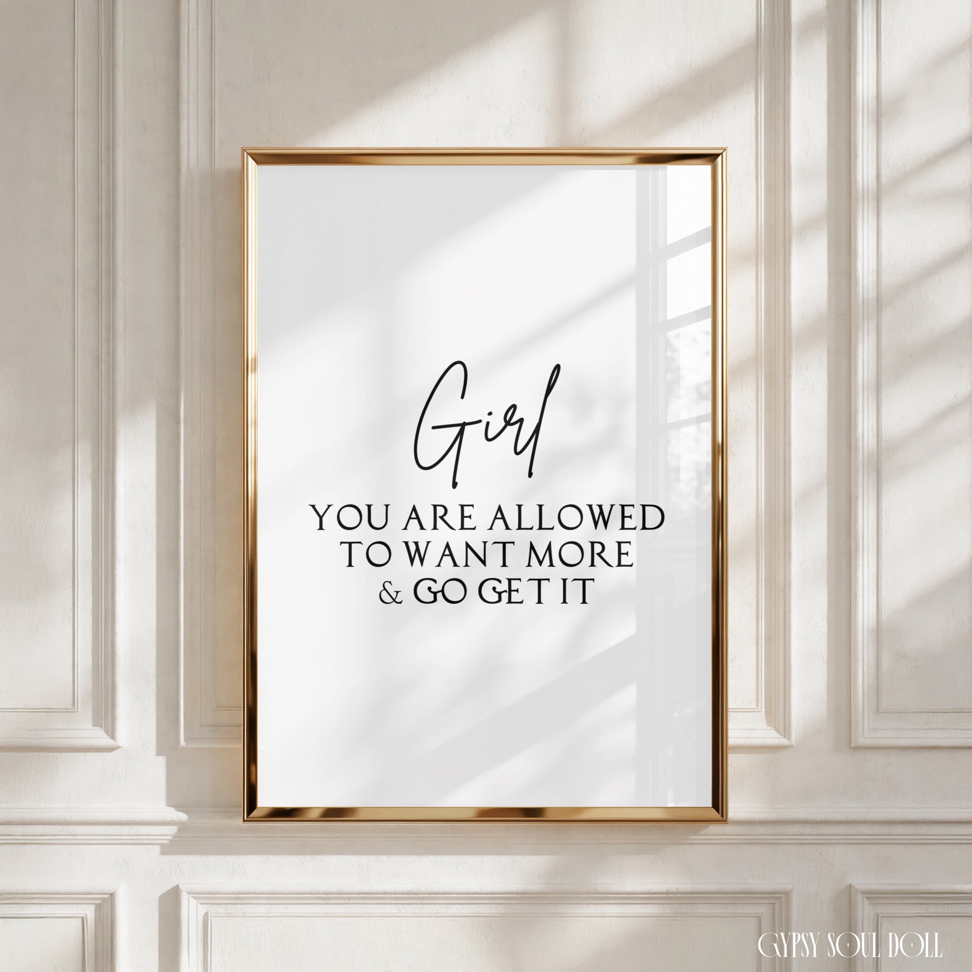 A framed typographic wall art print with a gold frame hangs on a white paneled wall. The print has black text reading “Girl YOU ARE ALLOWED TO WANT MORE & GO GET IT” on a white background.