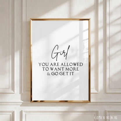 A framed typographic wall art print with a gold frame hangs on a white paneled wall. The print has black text reading “Girl YOU ARE ALLOWED TO WANT MORE & GO GET IT” on a white background.