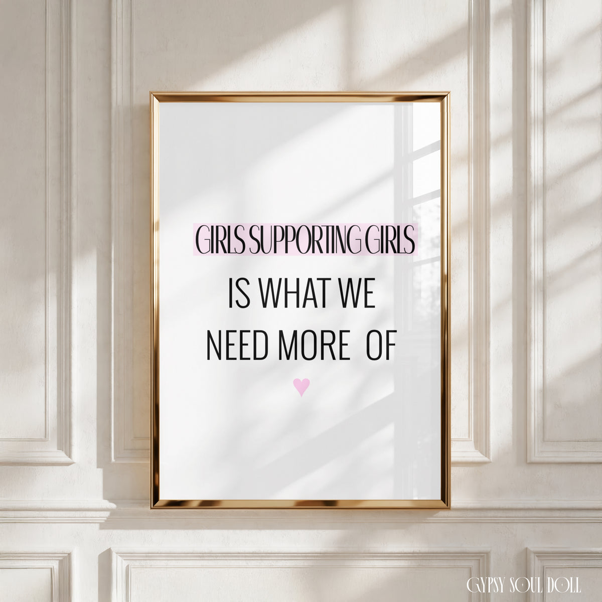 A framed typographic wall art print leaning against a white panelled wall. The print has a gold frame and reads, 'GIRLS SUPPORTING GIRLS IS WHAT WE NEED MORE OF' with a small pink heart below.